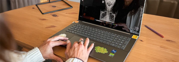 A person is using a laptop on a desk. The screen displays a medical image of a human skeleton, likely an X-ray. The laptop has colorful stickers on the keyboard area.