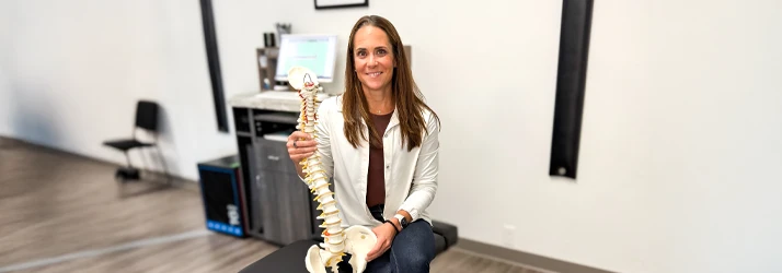 Dr. Sandy Ager seated in a clinical setting, holding a detailed anatomical spine model to illustrate spinal health and alignment.